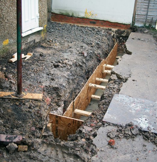 A dug trench near a house foundation with exposed concrete footings, surrounded by dirt and construction materials, suggesting ongoing building work.